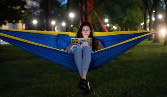 A woman sitting in a maize and blue hammock, looks at her laptop